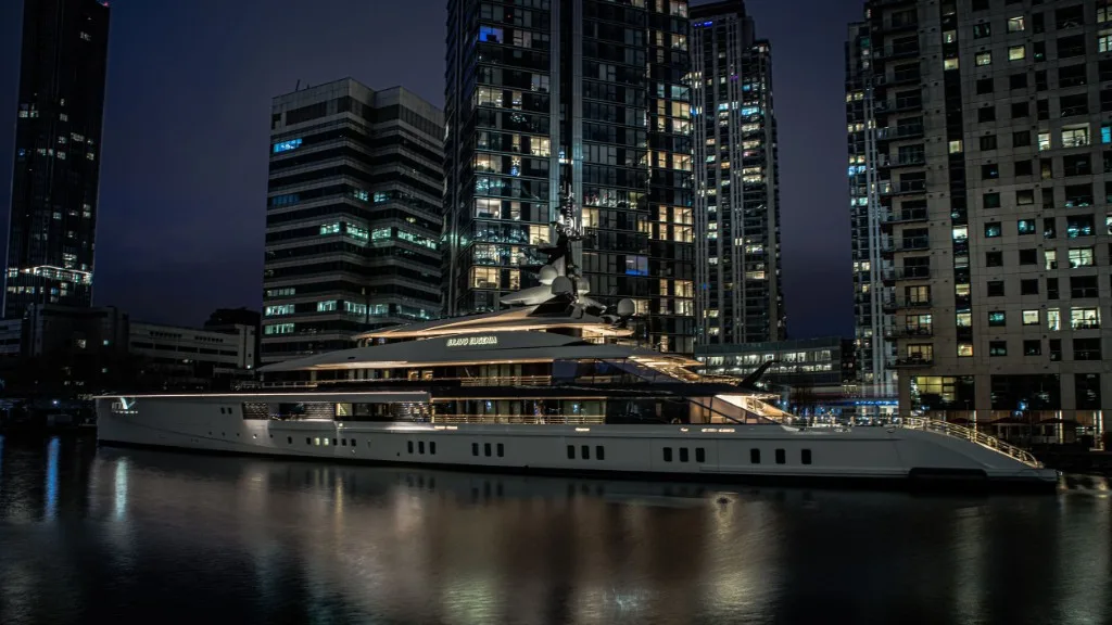 Luxury motor yacht Bravo Eugenia docked at night in a city harbor with illuminated skyscrapers behind it.