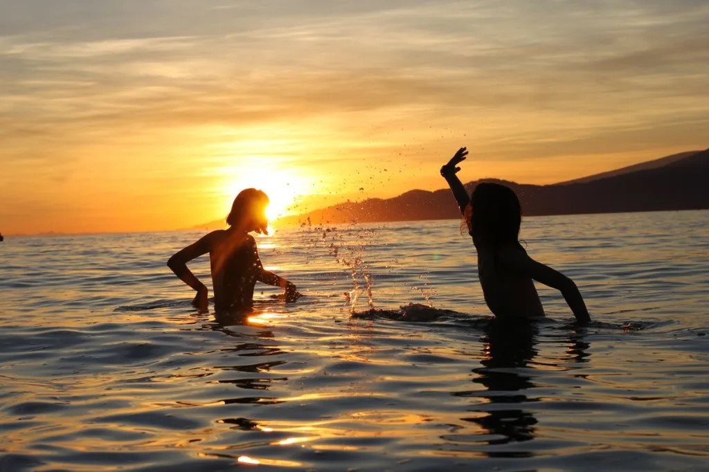 Two silhouetted figures playfully splashing in calm ocean water at sunset with a golden sky and distant coastline.