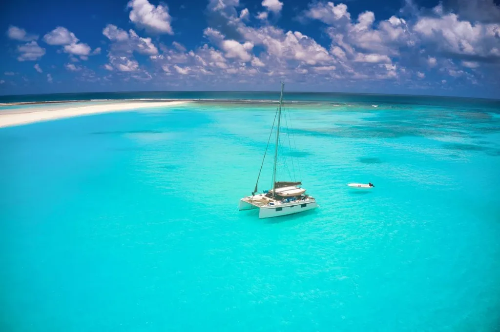 White luxury sailing catamaran anchored in clear turquoise tropical water beside a white sandbar under a blue sky.