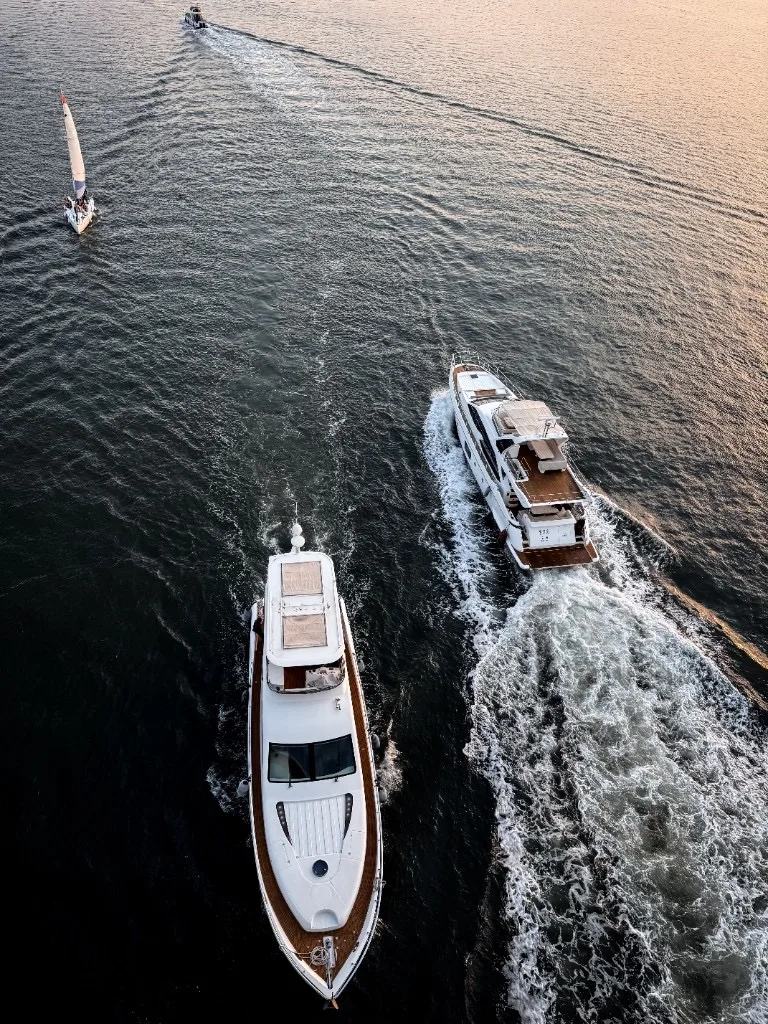 Aerial view of two white luxury motor yachts underway on dark blue water during golden hour, with teak decks and white wakes