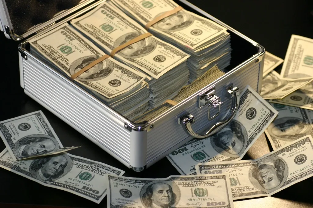 Open silver briefcase packed with stacked U.S. hundred-dollar bills, with loose bills scattered on a dark surface.