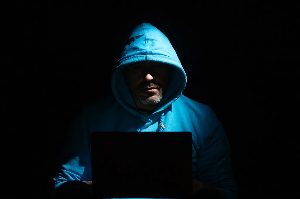 Person in a hooded sweatshirt lit by a laptop screen in a dark room, suggesting unauthorized access to tax accounts