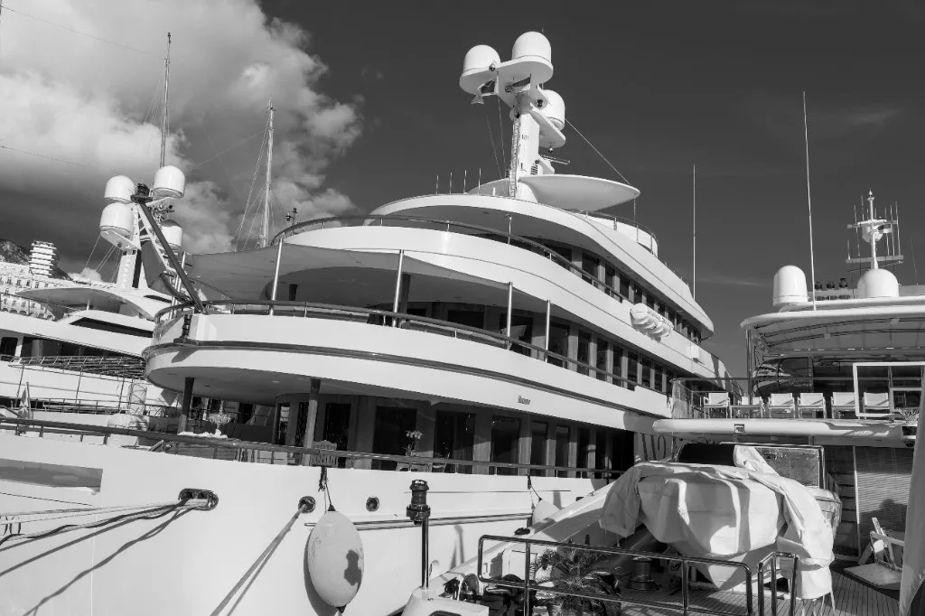 Black-and-white low-angle view of a multi-deck luxury motor yacht docked in a marina, with radar domes on the mast, mooring lines in the foreground, and a covered tender on a lower deck